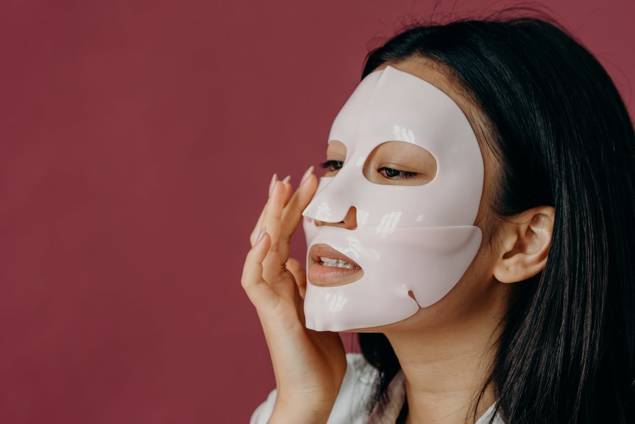 Asian woman applying a facial sheet mask against a red background, focusing on skincare and self-care.